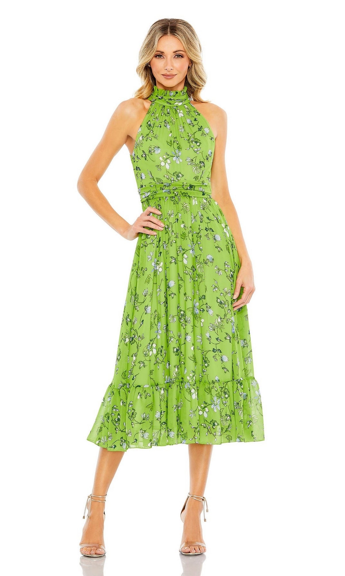 A woman wears the Short Wedding Guest Dress 55854 by Mac Duggal—a sage green, floral print, sleeveless halter-neck midi with a fitted waist—paired with beige high-heeled sandals, standing against a white background.